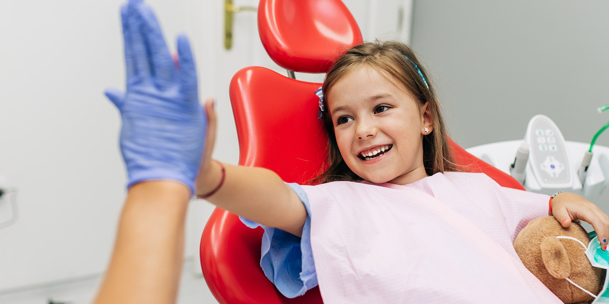 Cute kid in red dental chair holding a bear giving a high five to the dentist