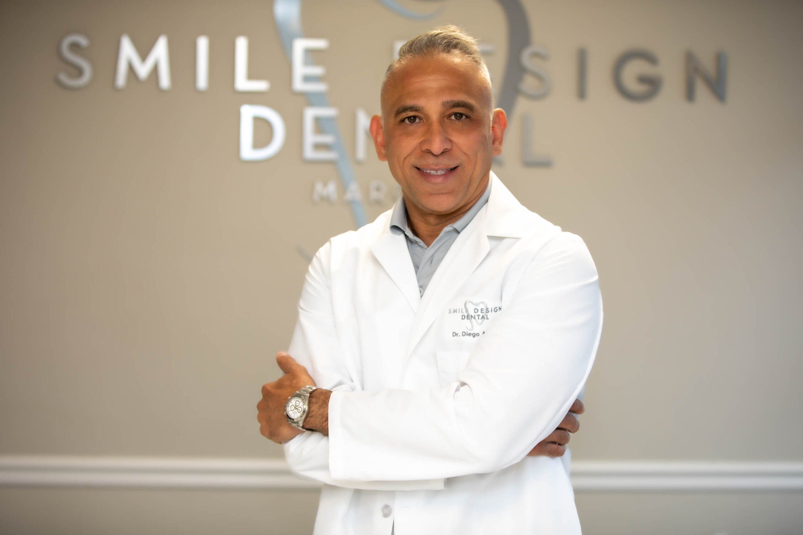 Dr. Azar standing professionally dressed and with his arms crossed across his chest in front of his practice name office logo before treating a patient with full mouth dental implants.
